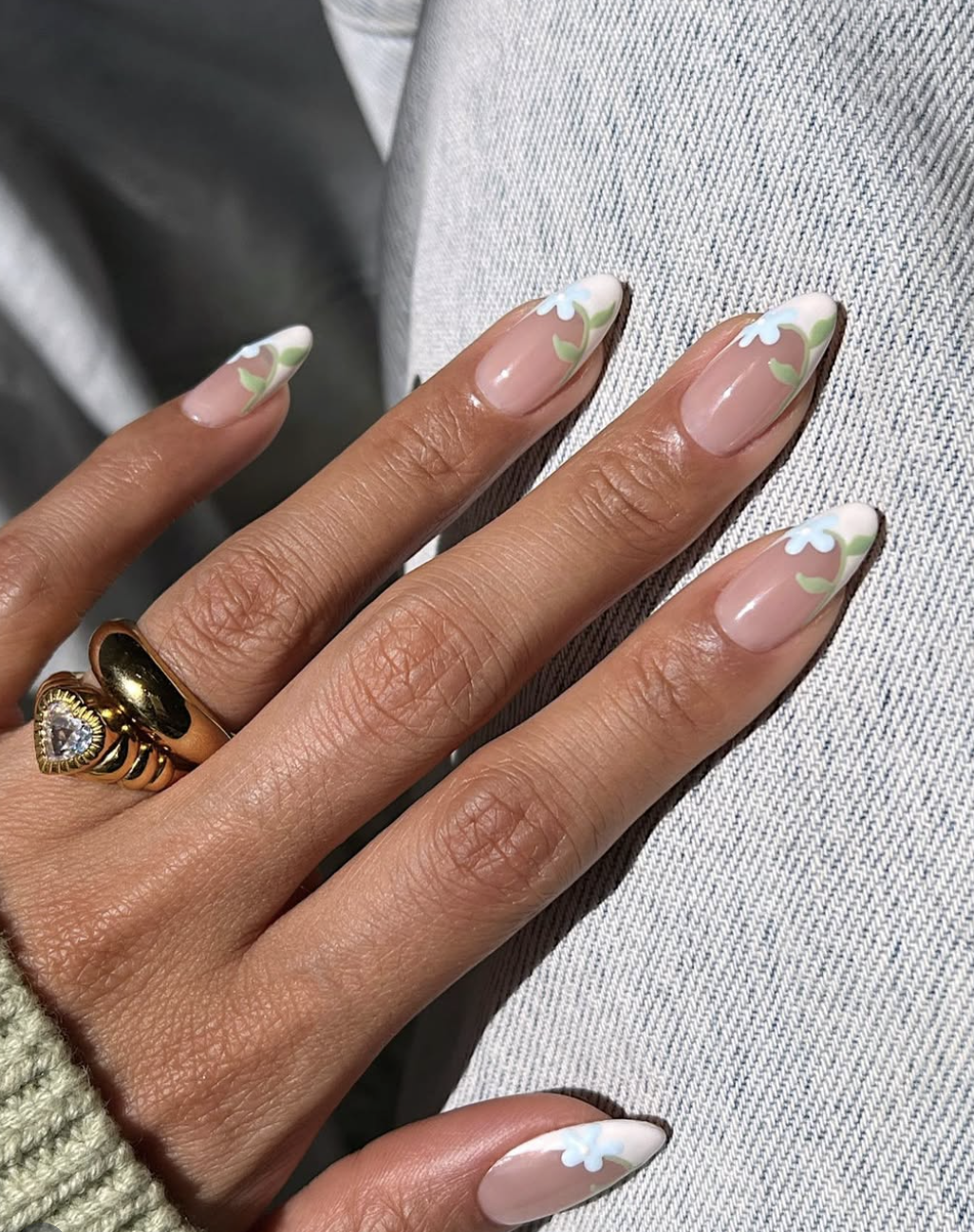 A close-up of a hand with medium almond-shaped nails featuring a sheer milky nude base and delicate hand-painted white flowers with green stems as French tips, styled with a gold heart-shaped ring.