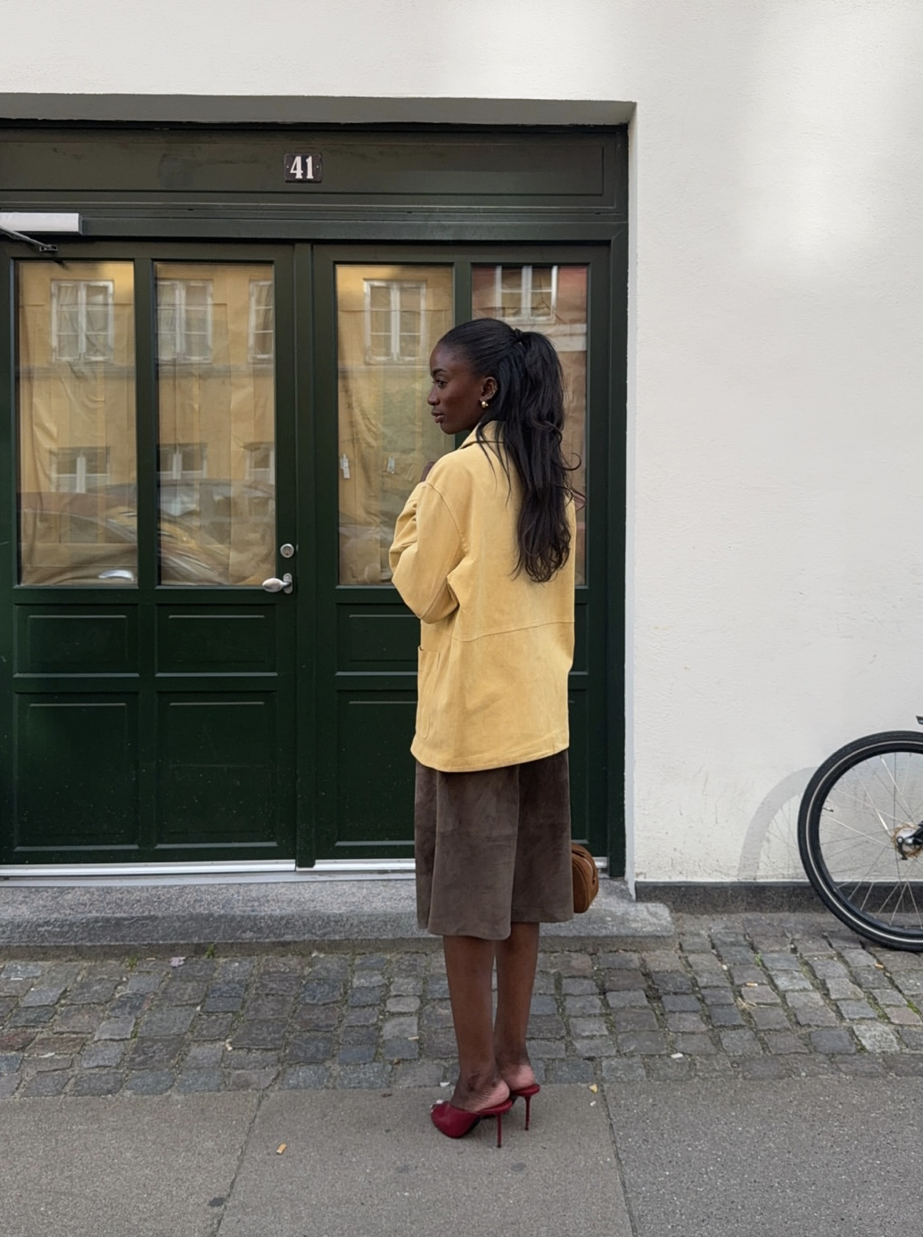 A woman with her hair in a half-up style wearing an oversized yellow jacket, a brown midi skirt, and dark red heeled mule sandals.