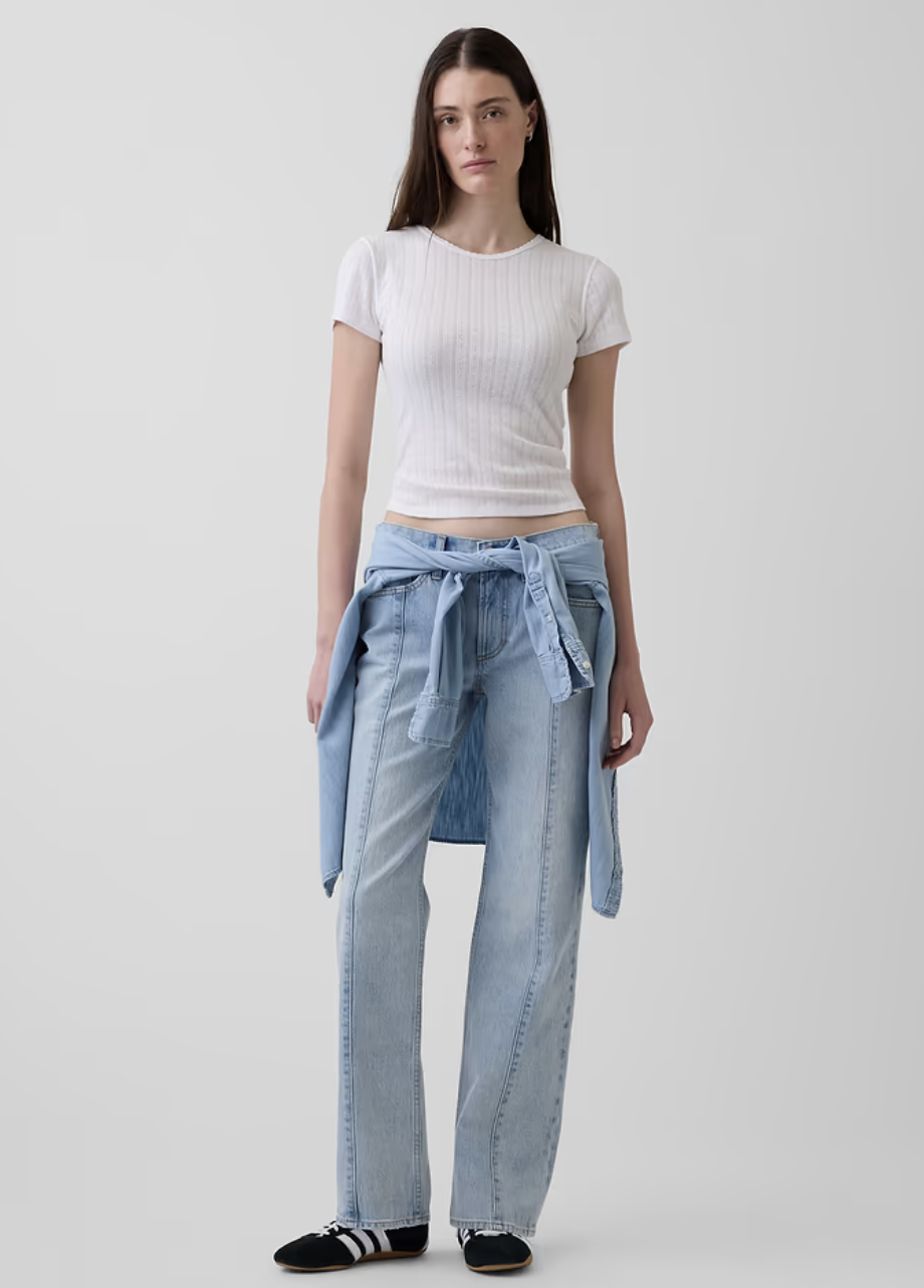 Model wearing relaxed light-wash jeans with a white t-shirt, light blue denim shirt tied at the waist, and black retro sneakers on a white background.