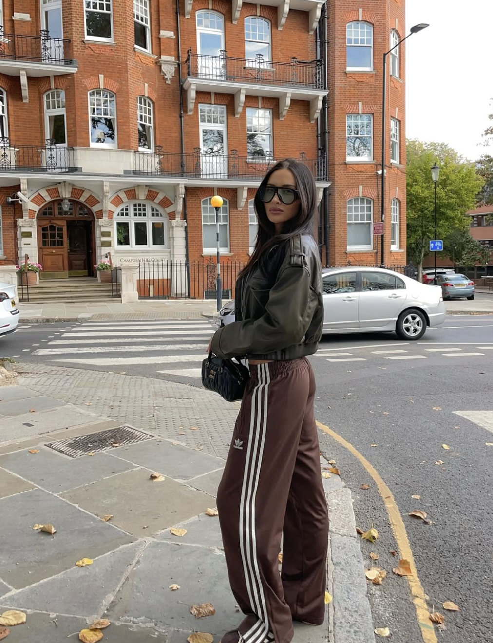 A woman wearing an elevated athleisure outfit featuring an oversized olive green bomber jacket, chocolate brown track pants, and a black structured shoulder bag.