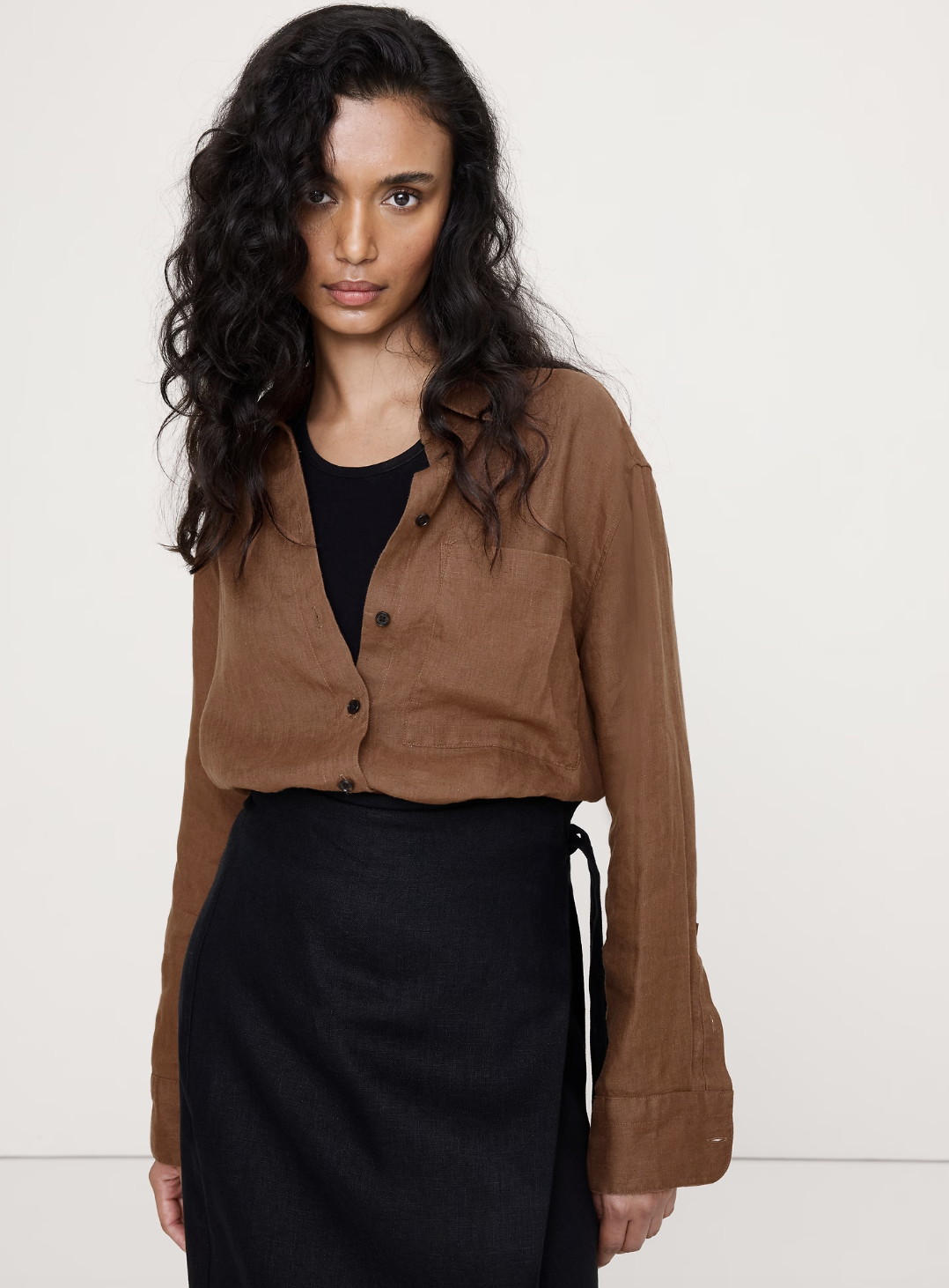 A professional work outfit featuring a brown button-down shirt tucked into a black midi wrap skirt, styled for a business casual look with sneakers.