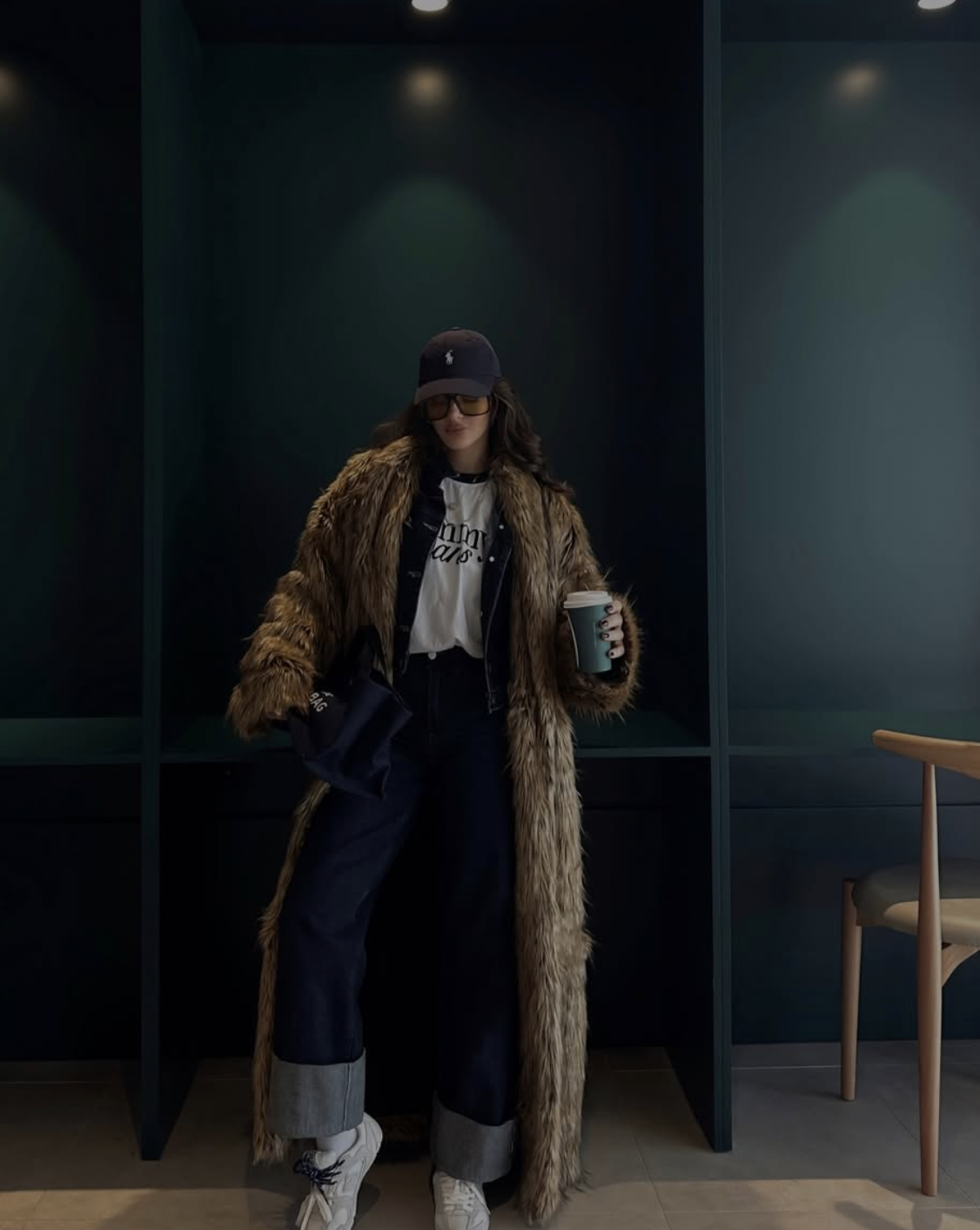 Woman wearing a long brown faux fur coat over a white graphic t-shirt and cuffed dark-wash jeans, paired with white sneakers and a navy baseball cap.