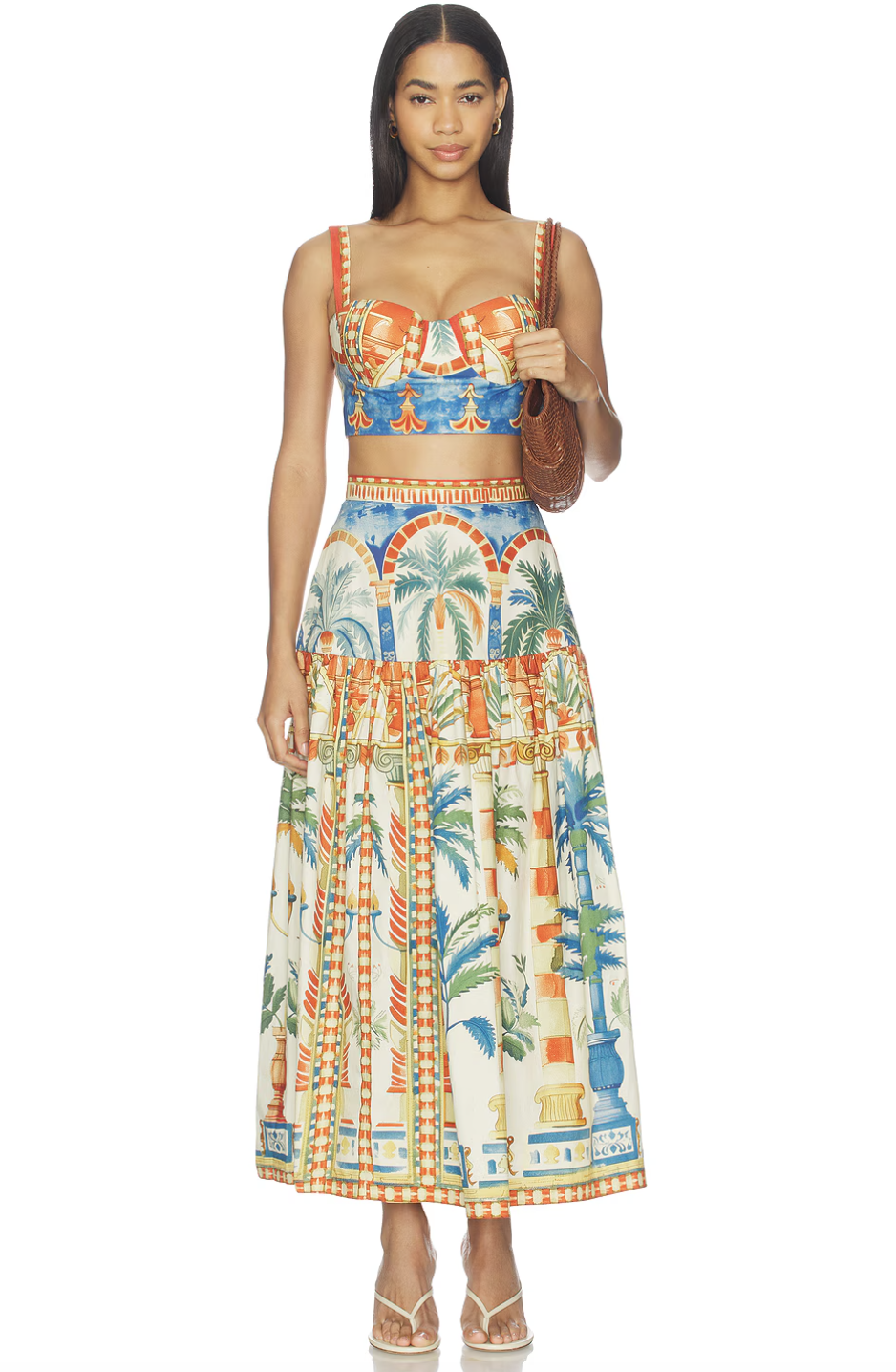 A woman wearing a colorful two-piece matching set featuring a tropical palm tree and architectural print, including a structured sweetheart crop top and a high-waisted tiered maxi skirt, while carrying a brown woven shoulder bag.