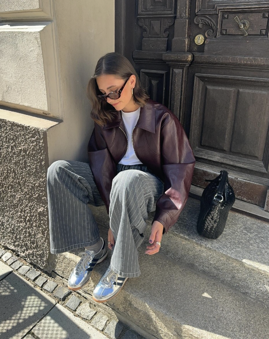 Woman sitting outdoors wearing an oversized burgundy faux leather jacket, grey wide-leg jeans, and metallic silver sneakers, styled with dark sunglasses.