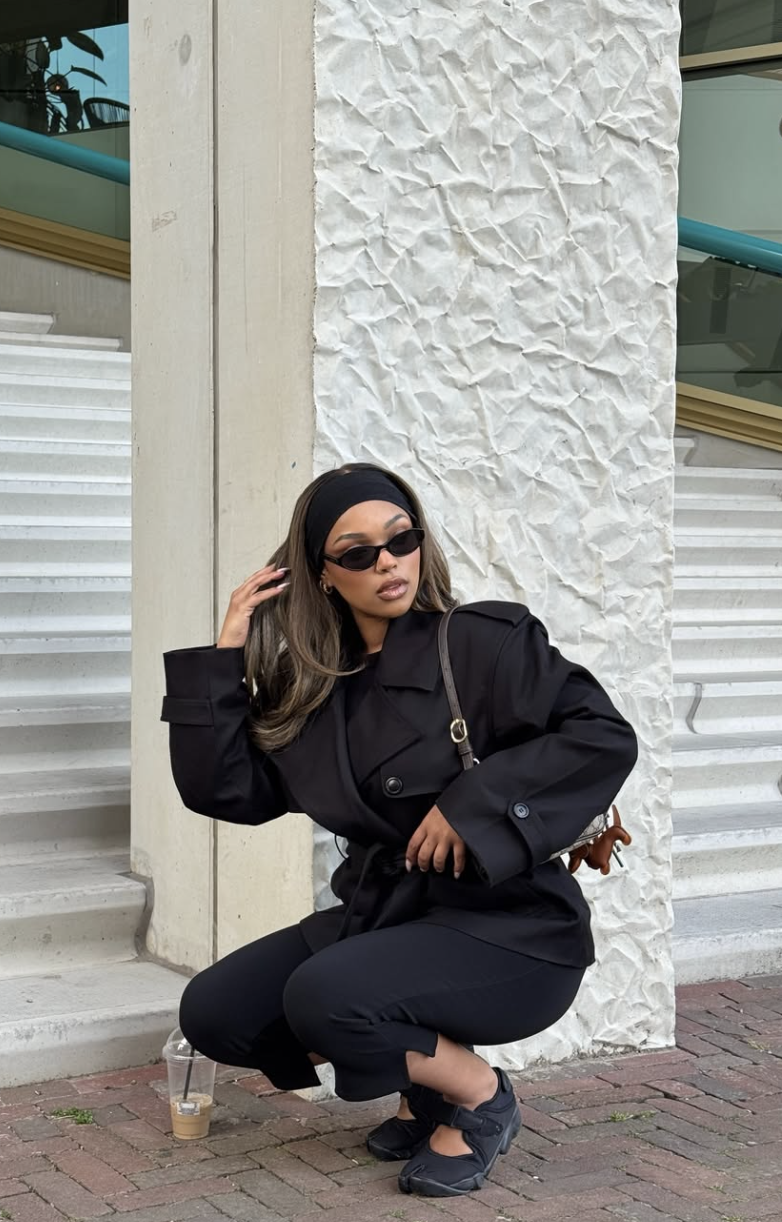Woman wearing a black cropped trench coat with wide sleeves and a belt, styled with a black headband and sunglasses while crouching outdoors.