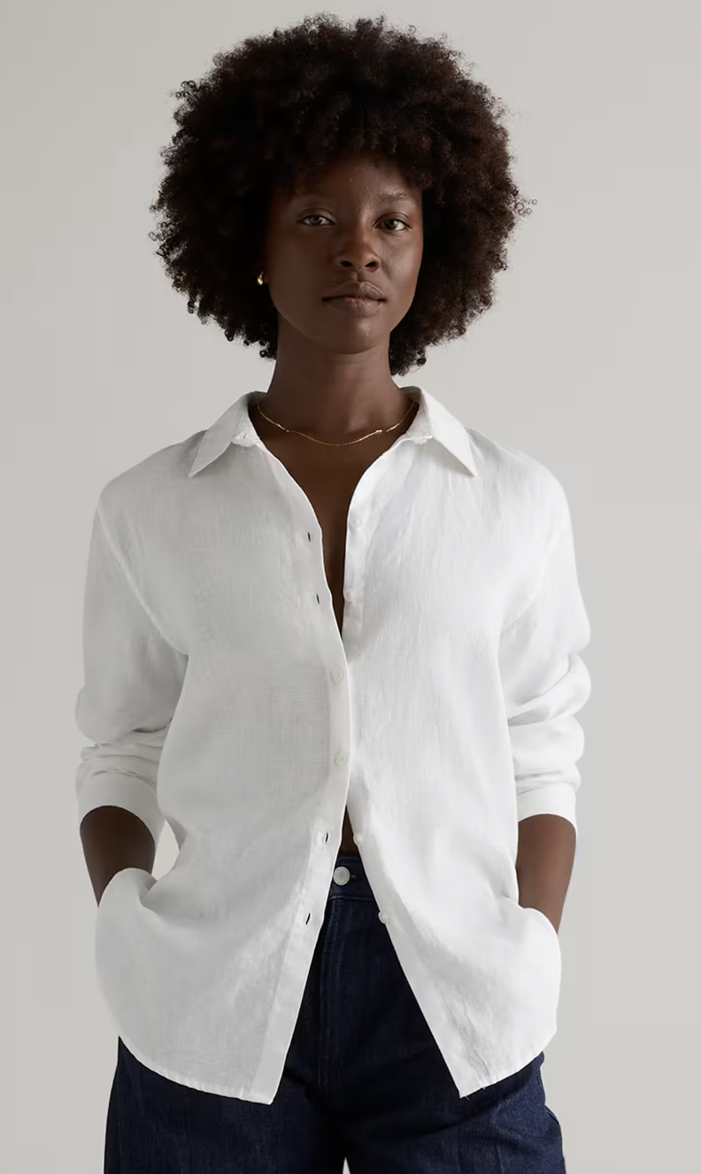 A close-up of a woman styling an oversized white linen button-down shirt paired with high-waisted dark denim jeans.