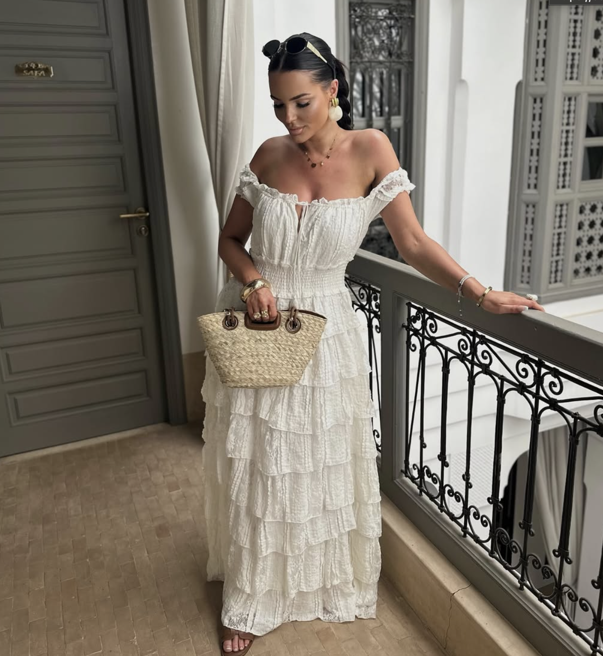 A woman styling a white off-the-shoulder smocked maxi dress with ruffled tiers, accessorized with a small woven straw handbag, chunky gold bracelets, a delicate gold necklace, and black sunglasses.