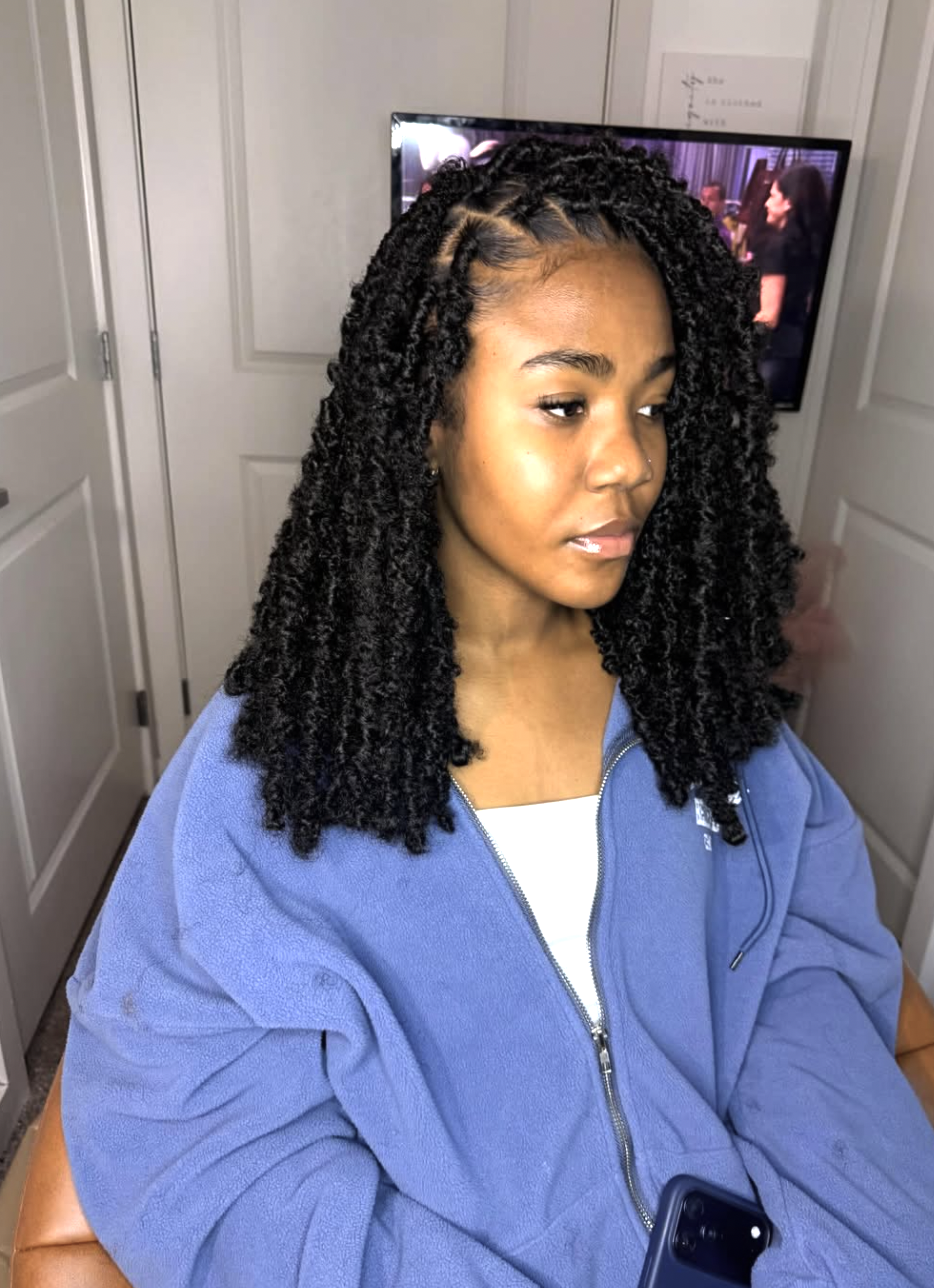 A woman showcasing shoulder-length butterfly locs with a distressed, bohemian texture and a clean side part, wearing a blue zip-up hoodie.