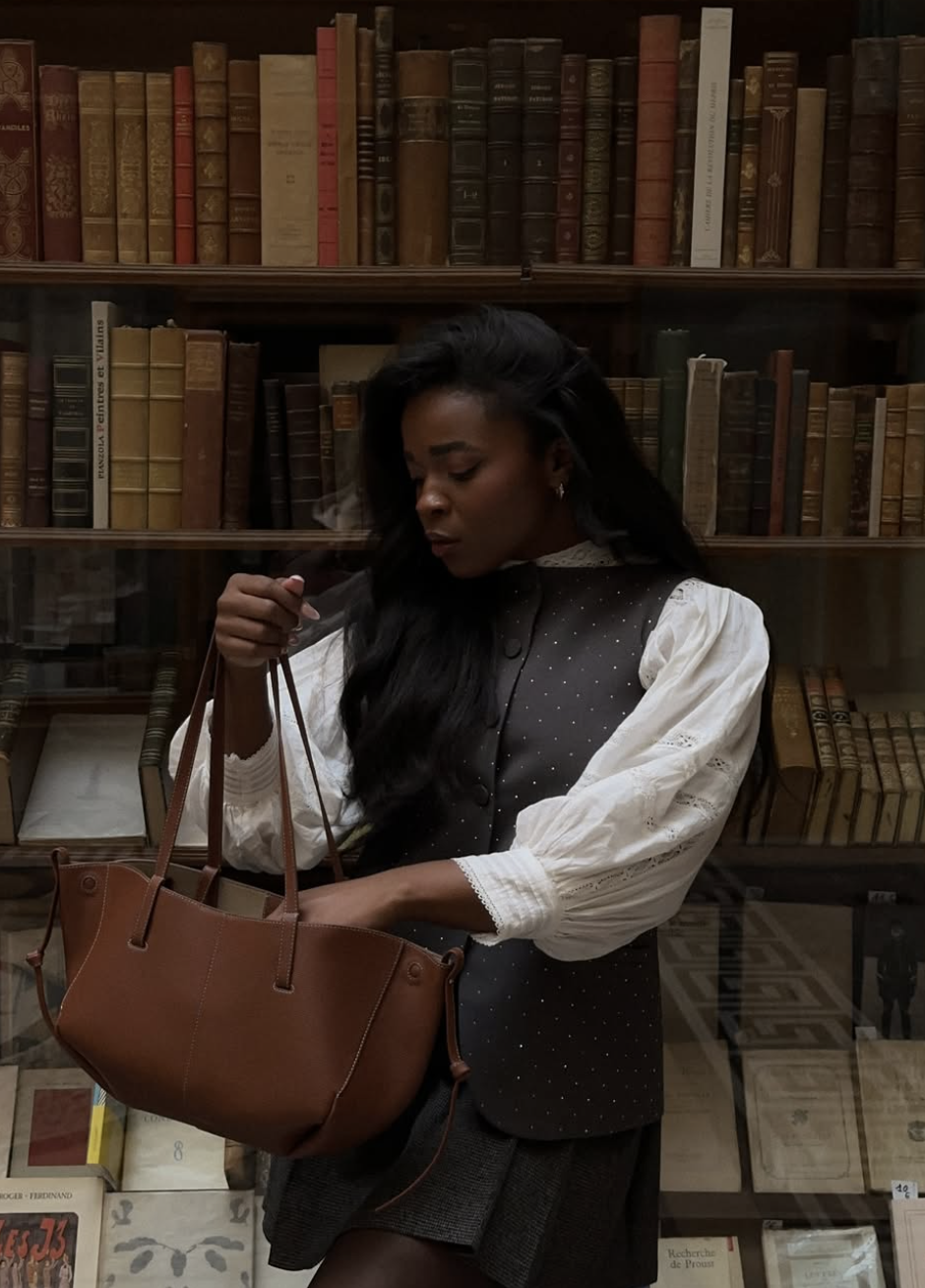 A woman in a dark vest and white poet blouse holding a brown leather satchel bag in a vintage library, capturing the poetcore aesthetic.