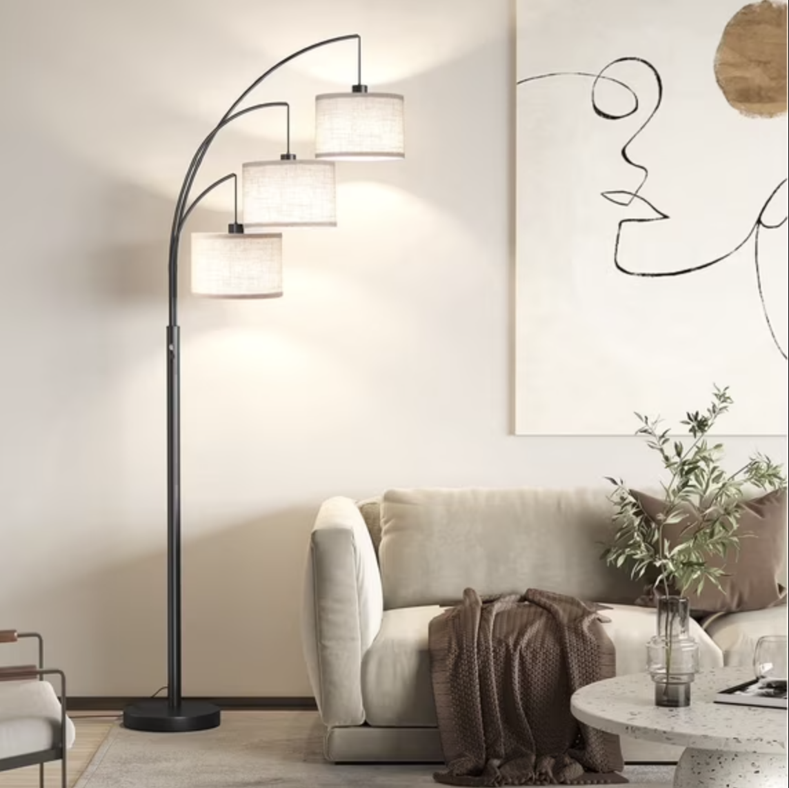 A modern black three-light arced floor lamp with textured drum shades positioned next to a beige sofa in a neutral living room.