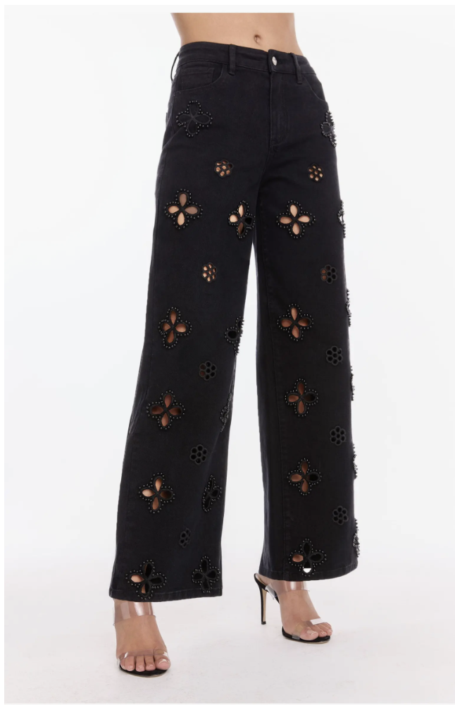A woman wearing black high-rise jeans from The Noli Shop featuring floral cutouts with pearl embellishments and a straight-leg fit.