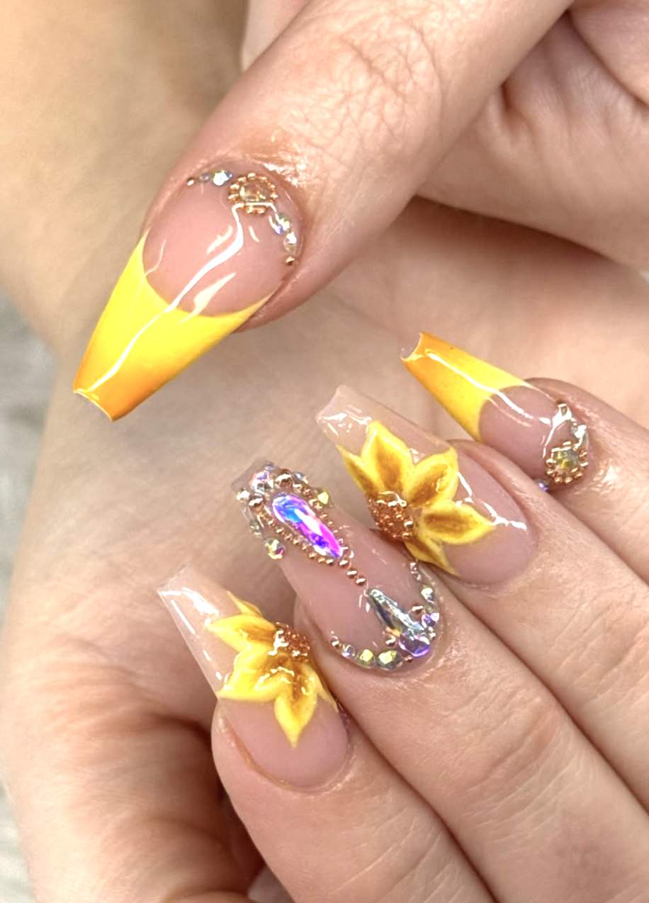 Long coffin-shaped nails with a bright yellow French tip, featuring 3D sunflower charms on clear accent nails with gold leaf and rhinestone details.