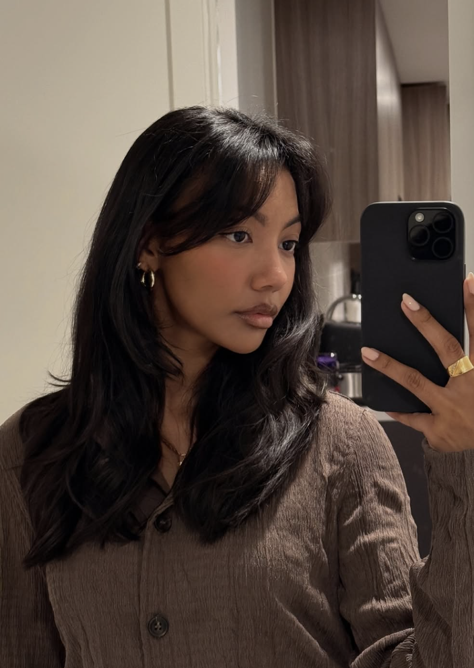 Asia Jackson wearing a brown button-up shirt and gold hoop earrings, featuring long dark hair with soft curtain bangs that frame the forehead to add definition to a round face shape.