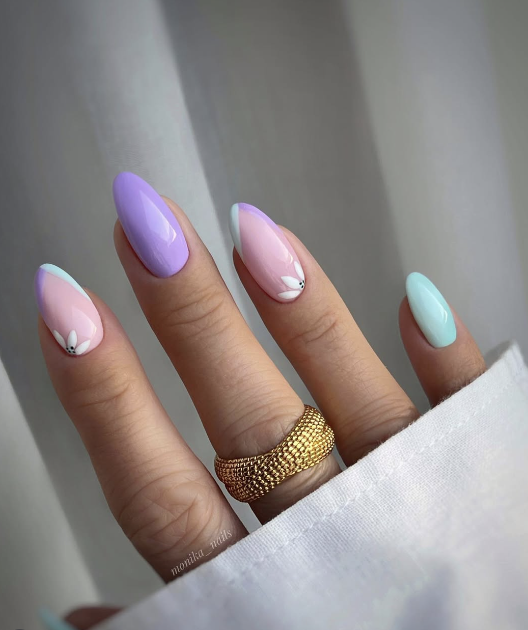 A close-up of a hand with long almond-shaped nails featuring a mismatched pastel design in lavender and soft pink, with white daisy floral accents and a chunky gold textured ring.