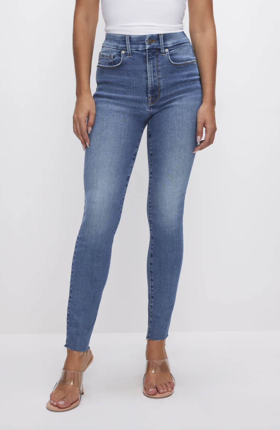 A woman wearing Good American Always Fits Skinny Jeans in a medium blue wash. The high-waisted denim is styled with clear strap heels, highlighting the gap-proof waistband and body-hugging fit."