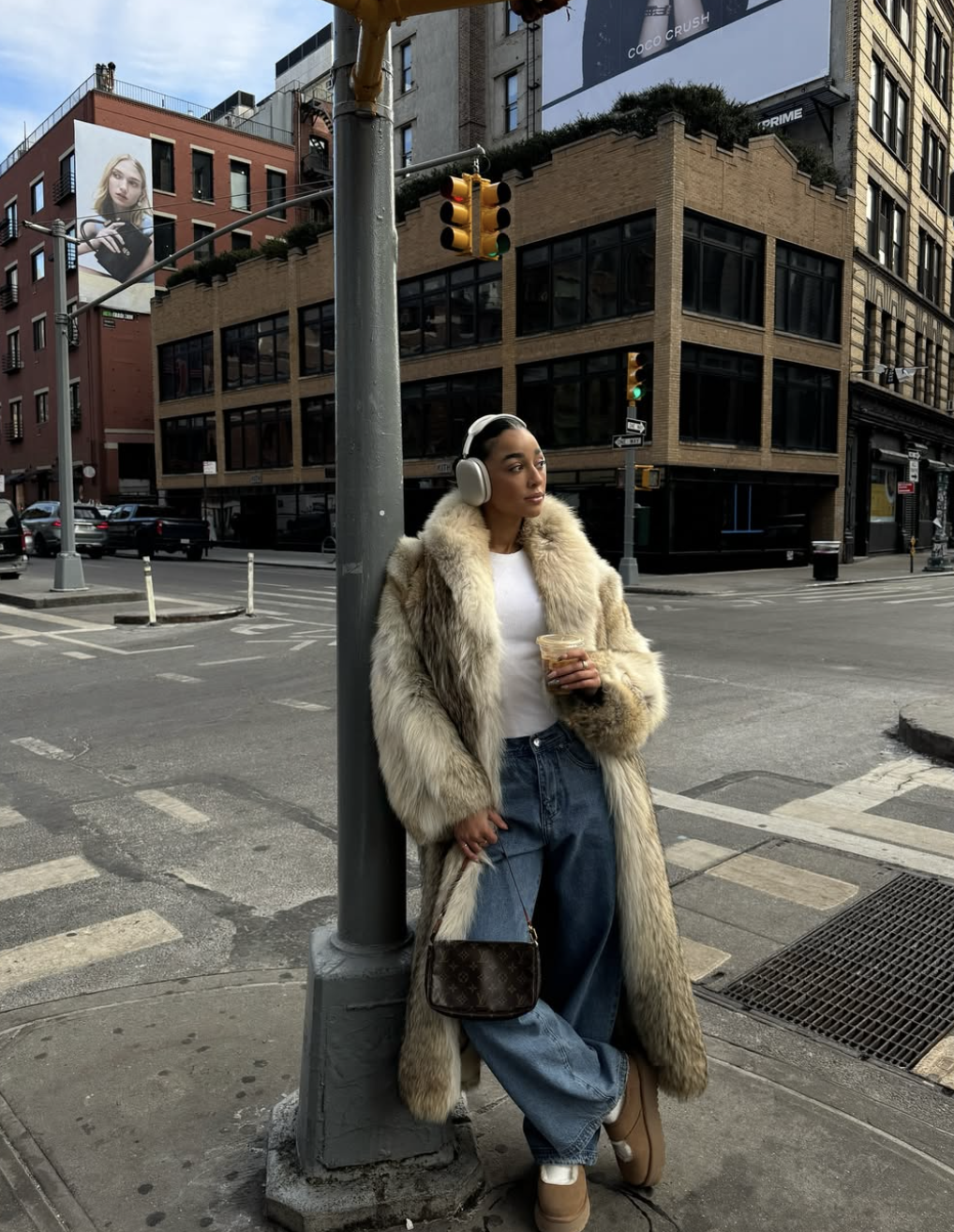 A woman leaning against a pole on a city street wearing a voluminous faux fur coat, baggy blue jeans, and black Mary Jane flats for women.