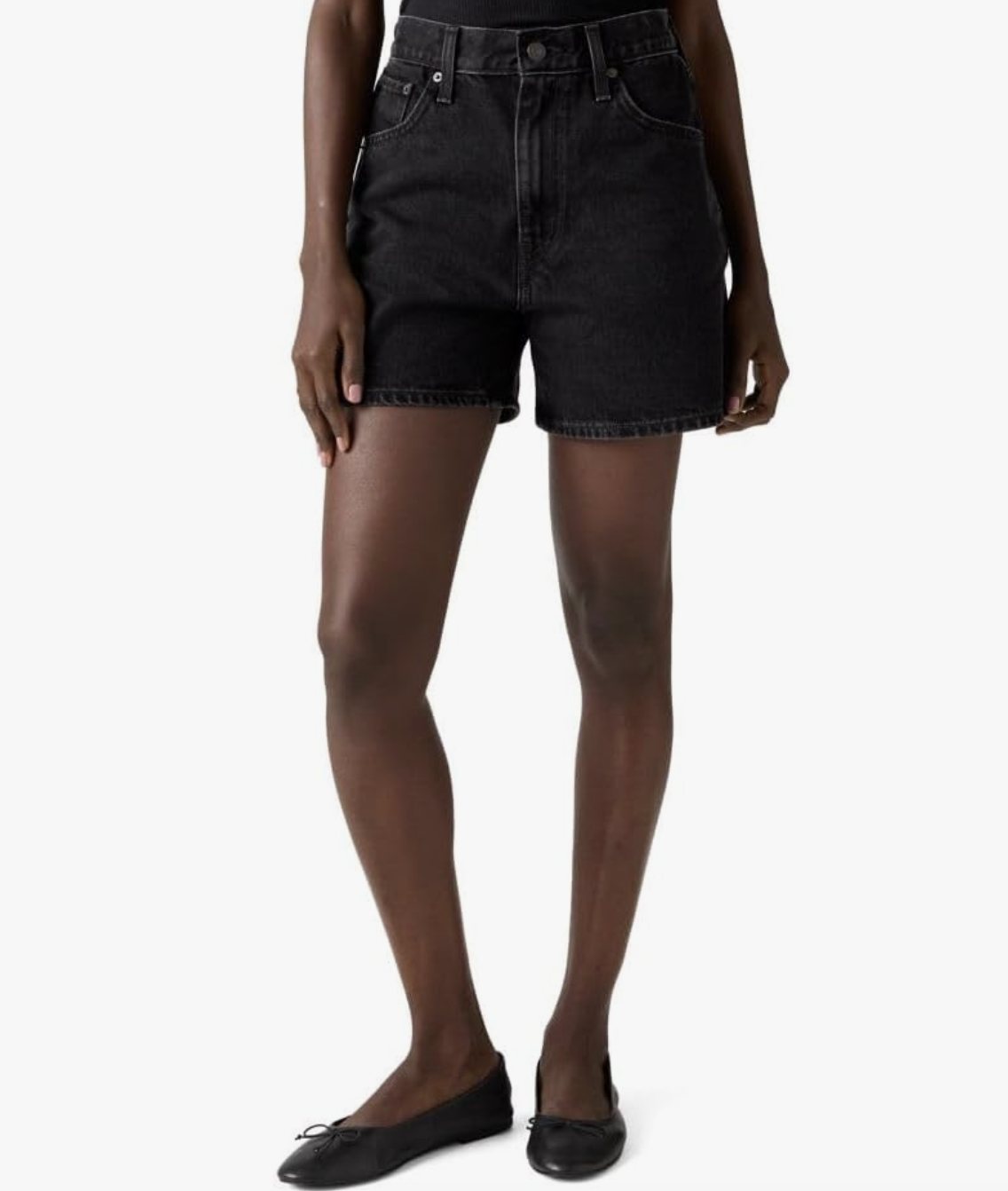 A close-up view of a woman's legs wearing relaxed black denim shorts and classic black leather ballet flats with a delicate bow detail.