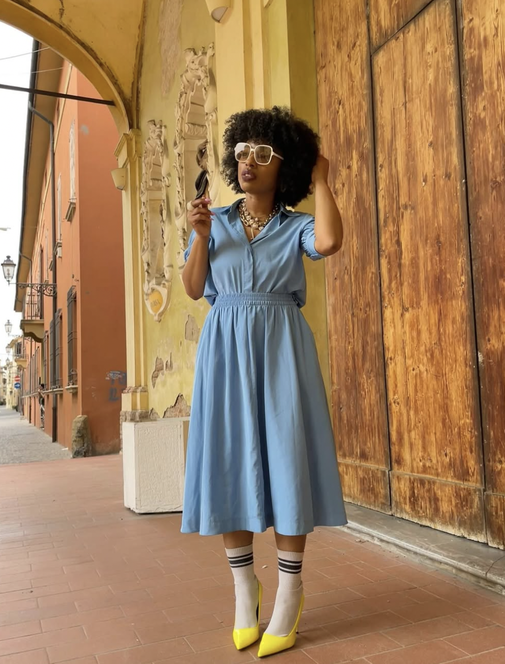 A woman with an afro hairstyle wearing a light blue midi dress, white socks with black stripes, and bright yellow pointed-toe high heels, standing on a brick walkway.