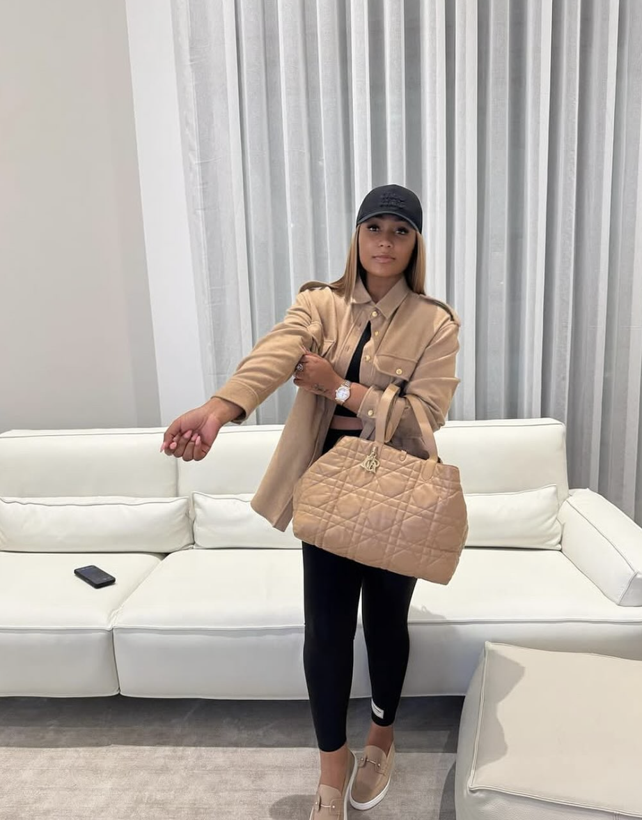 Influencer Muriel Blanche wearing black leggings with a tan button-down shirt jacket, a black baseball cap, and classic black loafers with gold hardware.