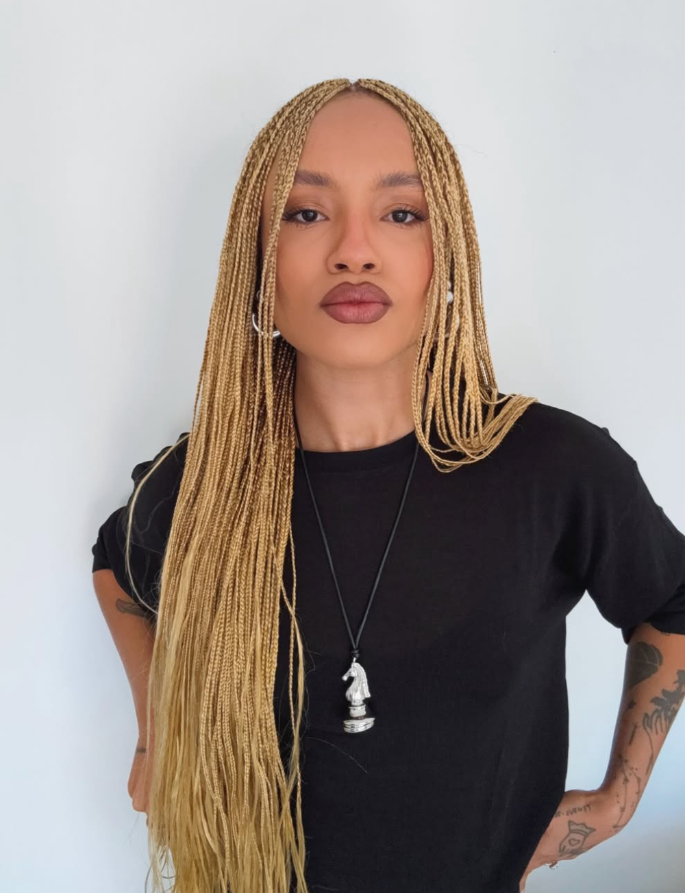 Close-up portrait of a woman showcasing long blonde micro braids styled with a clean middle part, wearing a black t-shirt and a silver horse pendant necklace.