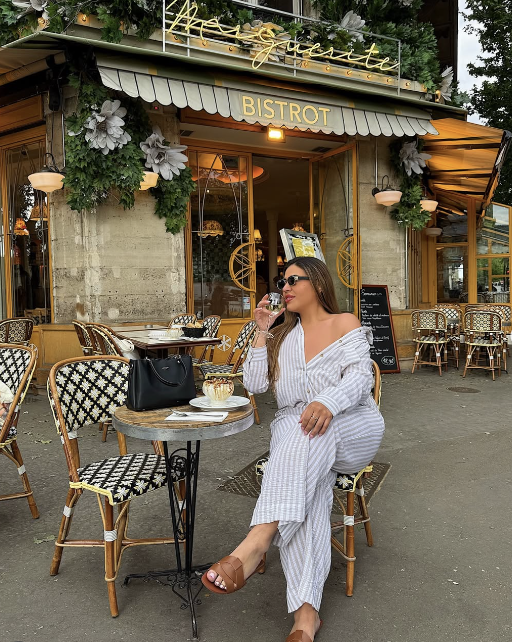 Plus-size woman sitting at an outdoor café table wearing a light gray and white striped matching button-down shirt and wide-leg pants set.