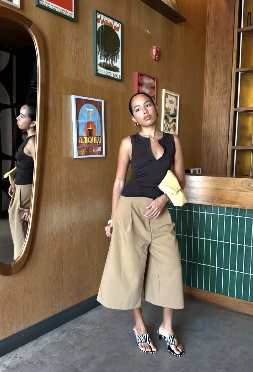 A woman wearing a black tank top and high-waisted, pleated beige wide-leg trousers styled with zebra-print mule sandals, standing in front of a mirror.