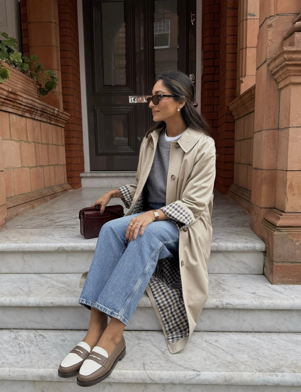 A woman wearing a tan longline trench coat layered over a striped sweater and straight-leg jeans with two-tone loafers.