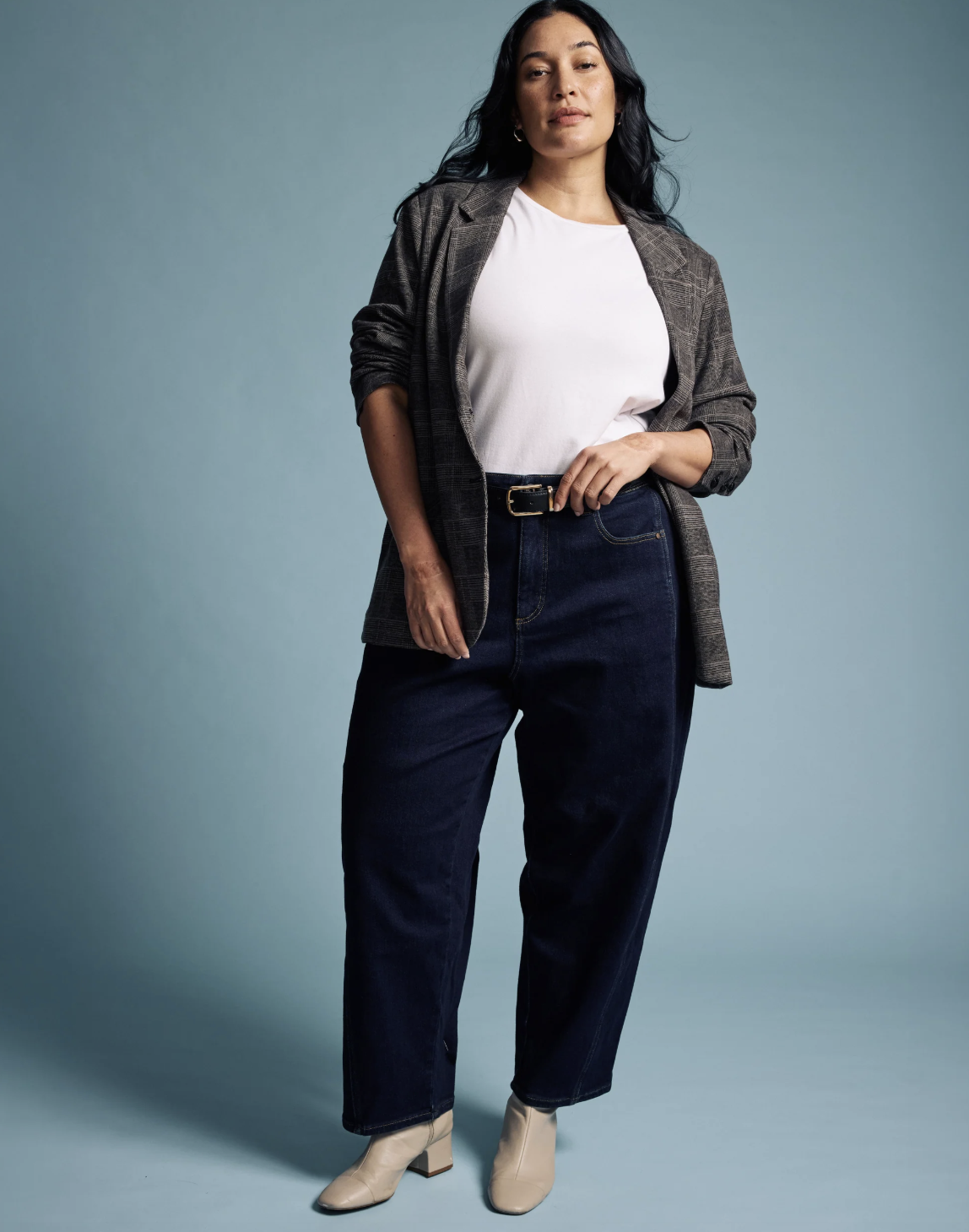 A person wearing dark indigo Universal Standard Dakota Twisted Leg Jeans, featuring a high-rise waist and a unique vertical twisted seam, styled with a white tee, grey blazer, and cream boots.