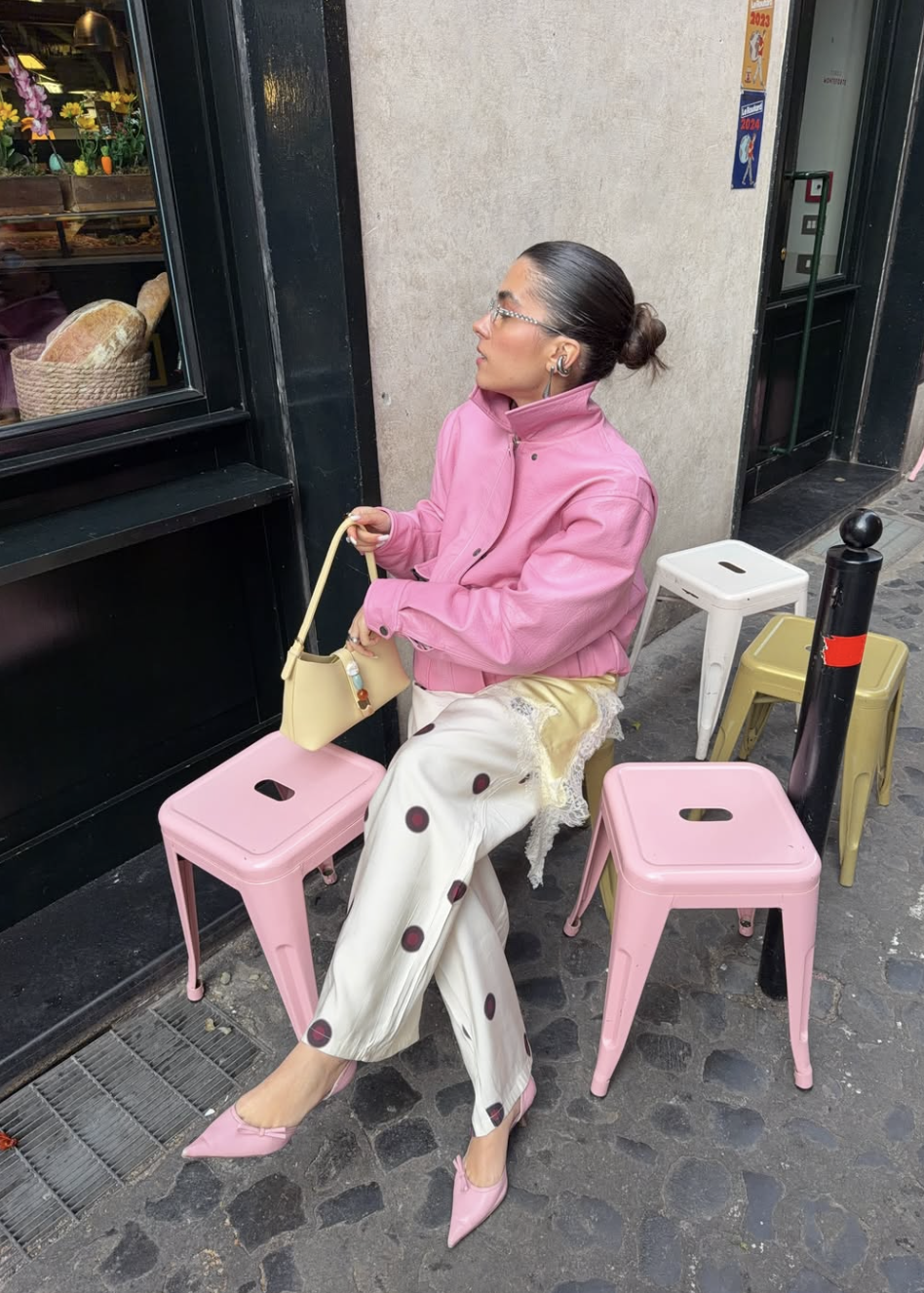 A woman wearing a bright pink zip-up jacket and white wide-leg trousers with large dark polka dots, holding a cream handbag.
