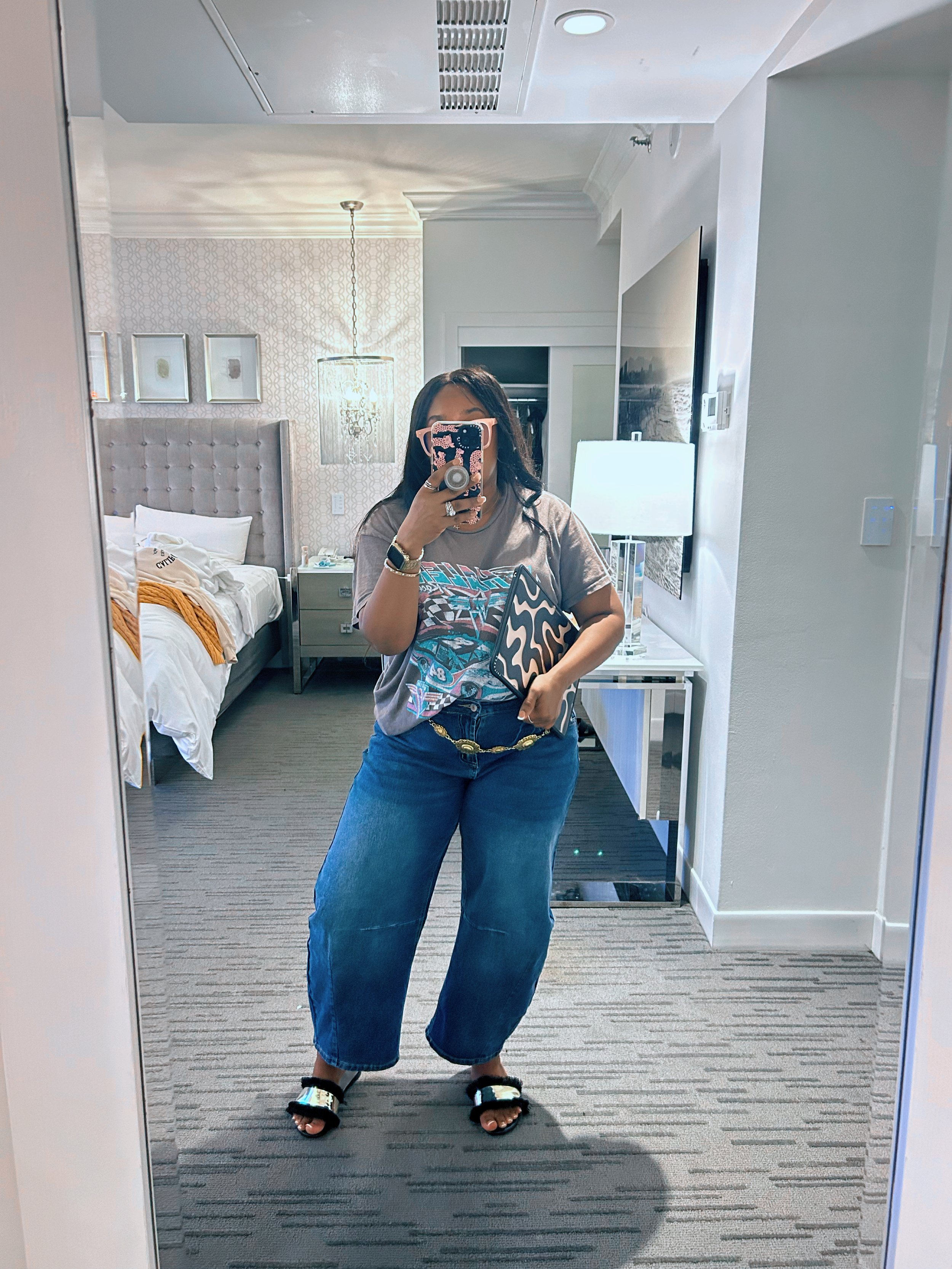 Woman wearing a gray graphic t-shirt partially tucked into high-waisted blue barrel jeans with a gold chain belt and black slide sandals.