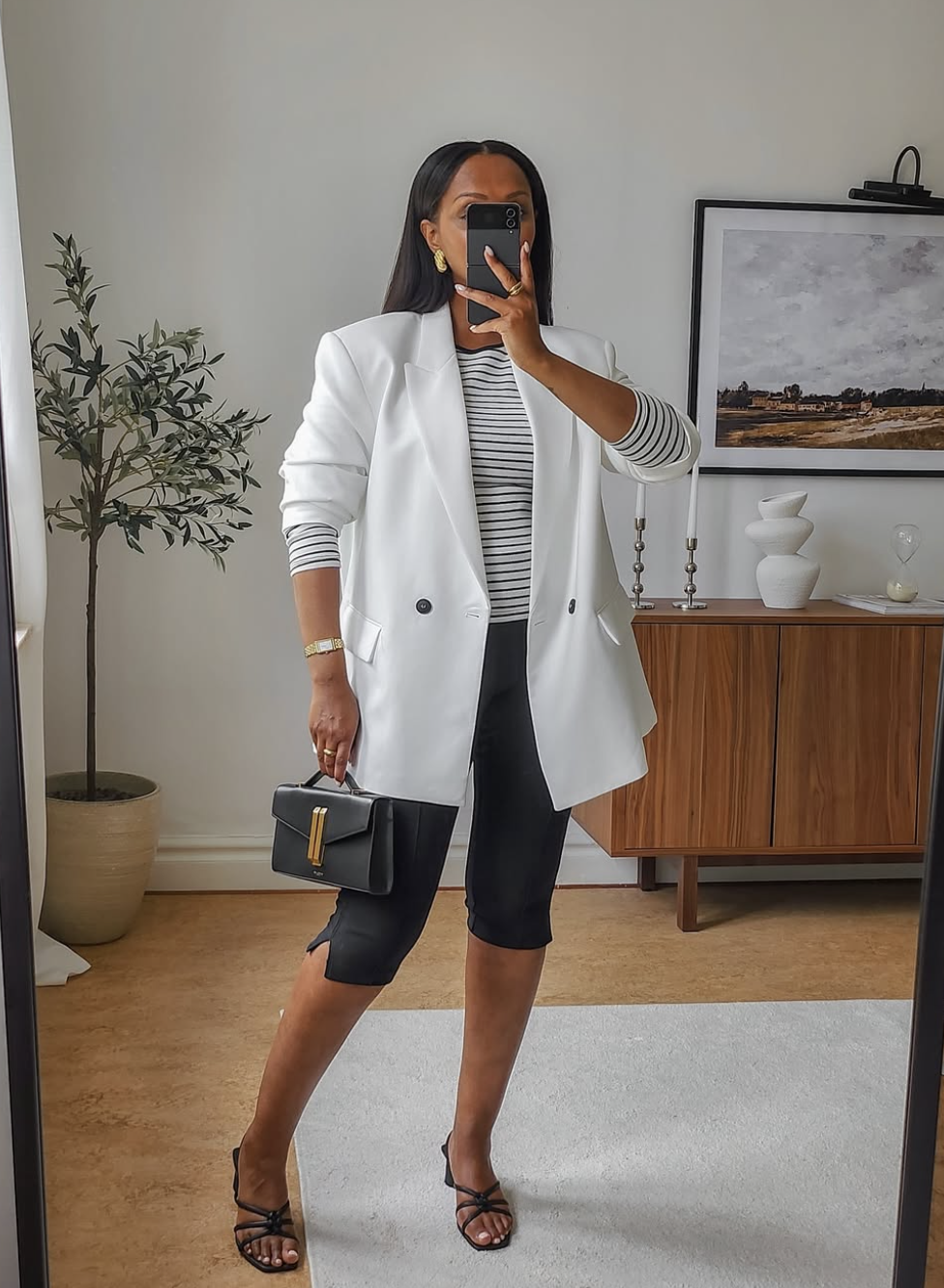 A woman wearing an oversized white blazer over a black and white striped t-shirt and black biker shorts, accessorized with a black structured handbag and strappy sandals.
