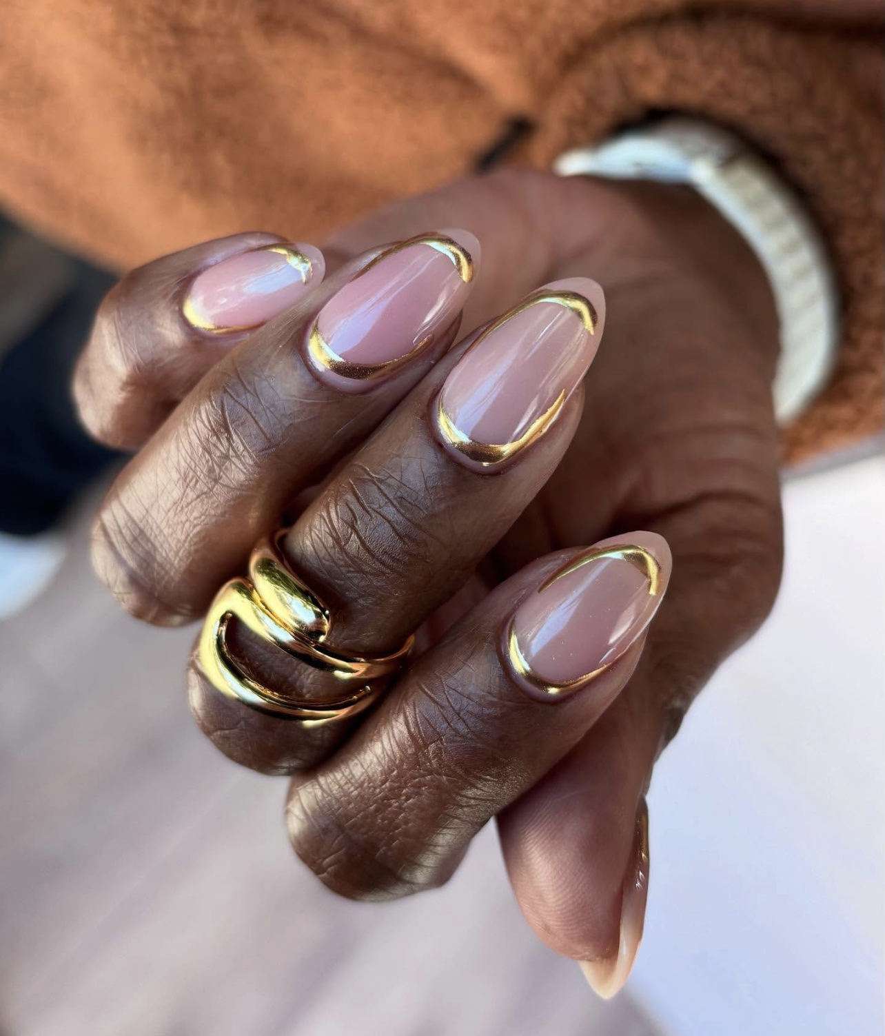 A close-up of almond-shaped nails featuring a sheer pink base with a highly reflective, metallic gold French outline tracing the edges, accessorized with a bold, chunky gold ring.