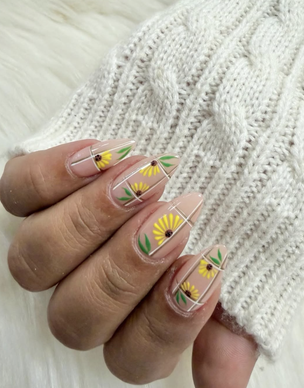 Almond-shaped nails with a nude base, featuring hand-painted sunflowers with green leaves and crisp white vertical and horizontal geometric lines.