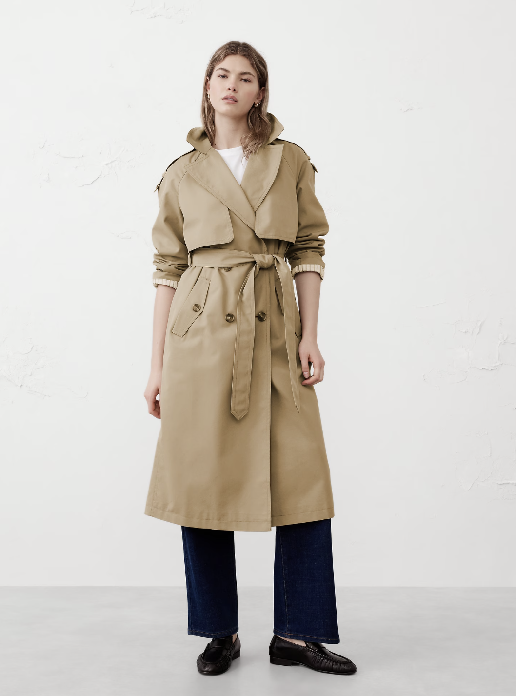 A woman wearing the tan Banana Republic Timeless Trench Coat with the belt tied in a front bow, styled with dark-wash wide-leg denim and black leather loafers.
