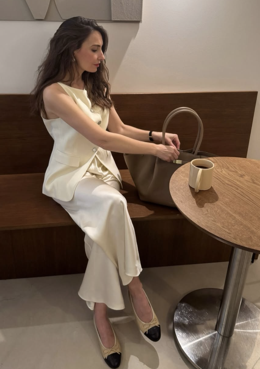 Petite woman sitting on a bench wearing a cream tailored vest and matching trousers paired with black pointed-toe ballet flats.