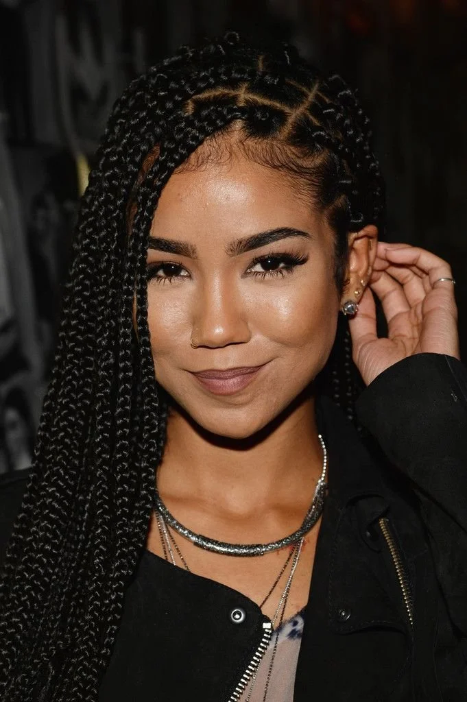 Jhené Aiko wearing classic waist-length box braids with a clean rectangular parting, paired with a silver chain necklace and a black jacket.