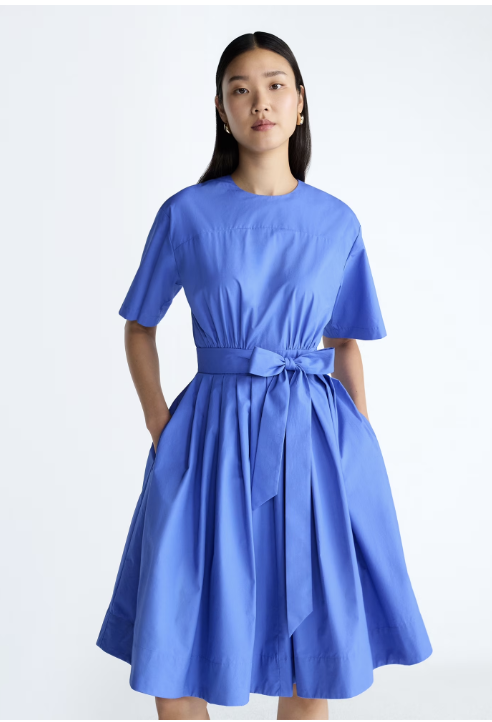 A woman wearing a bright blue short-sleeve midi dress with a structured cotton fabric and a tie-waist detail from the Scoop x Devil Wears Prada collection.