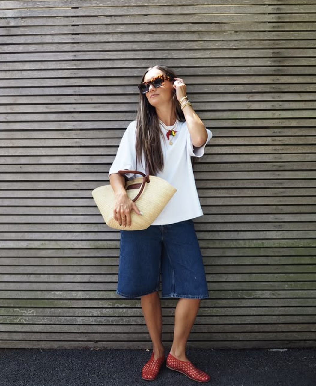 A street style look featuring a woman wearing relaxed, dark-wash denim Bermuda shorts, a classic white short-sleeve top, a baseball cap, a large woven straw tote bag, and bright red woven lattice jelly flat shoes.
