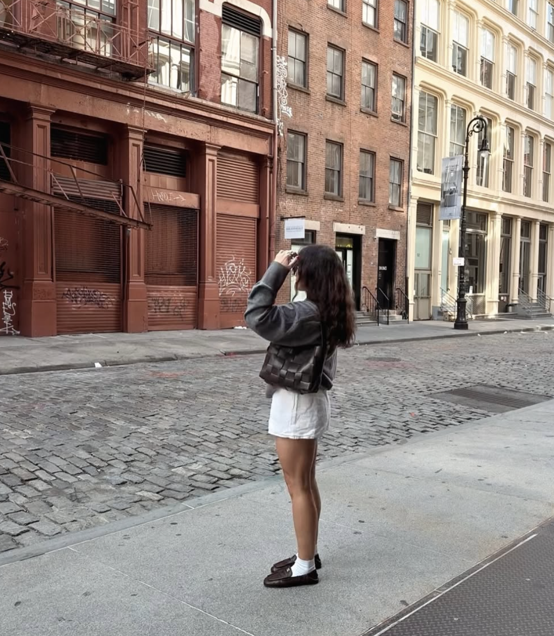 Woman standing on a cobblestone street wearing an oversized black faux leather jacket, a white mini skirt, and dark brown loafers with white socks.