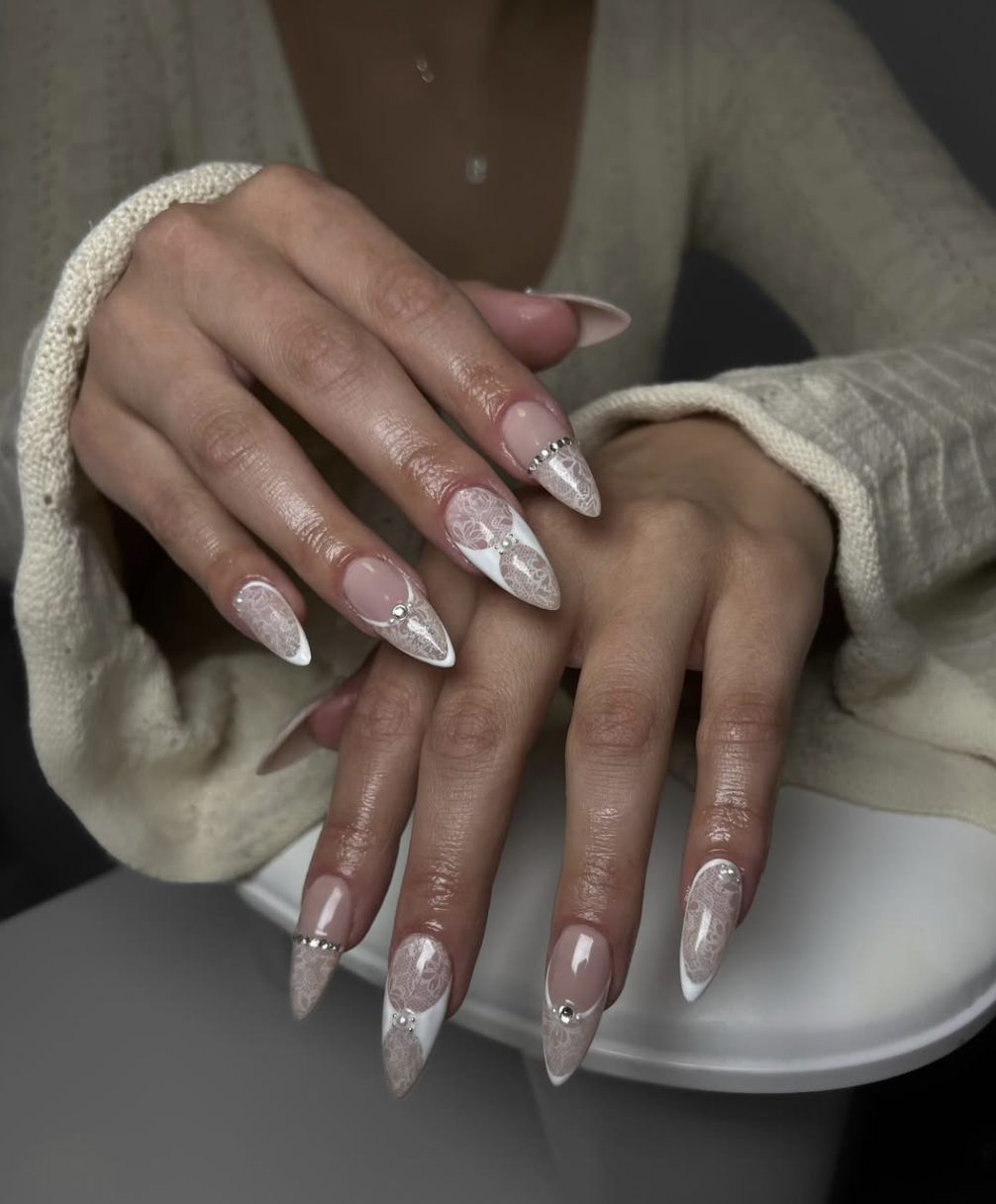 Long almond acrylic nails with a sheer nude base featuring white hand-painted lace patterns and 3D metallic gold wire accents.