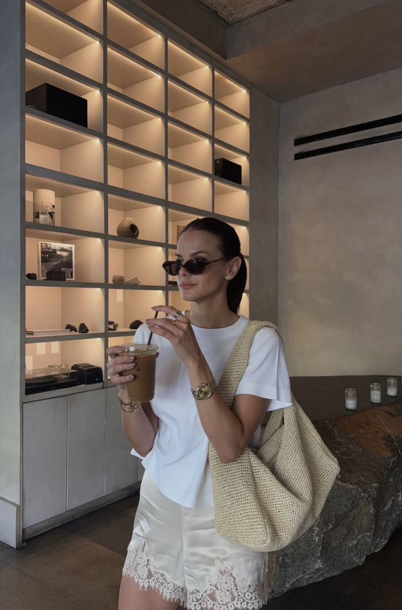 A woman wearing champagne-colored satin shorts with a lace hem, styled with a tucked-in white fitted t-shirt, a woven shoulder bag, and sunglasses.