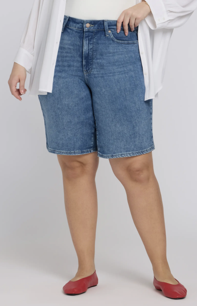 A woman wearing dark-wash NYDJ Lilly high-waisted denim shorts with a cuffed hem, designed with a wide leg opening to prevent riding up on thick thighs.