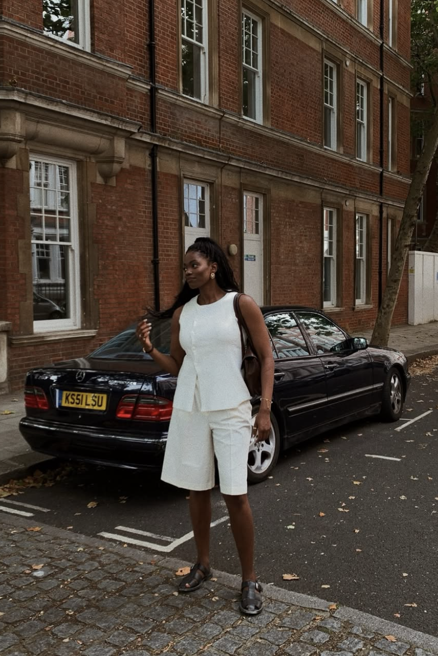 A woman wearing a white sleeveless vest and matching white Bermuda shorts, paired with black caged fisherman sandals and a black shoulder bag.