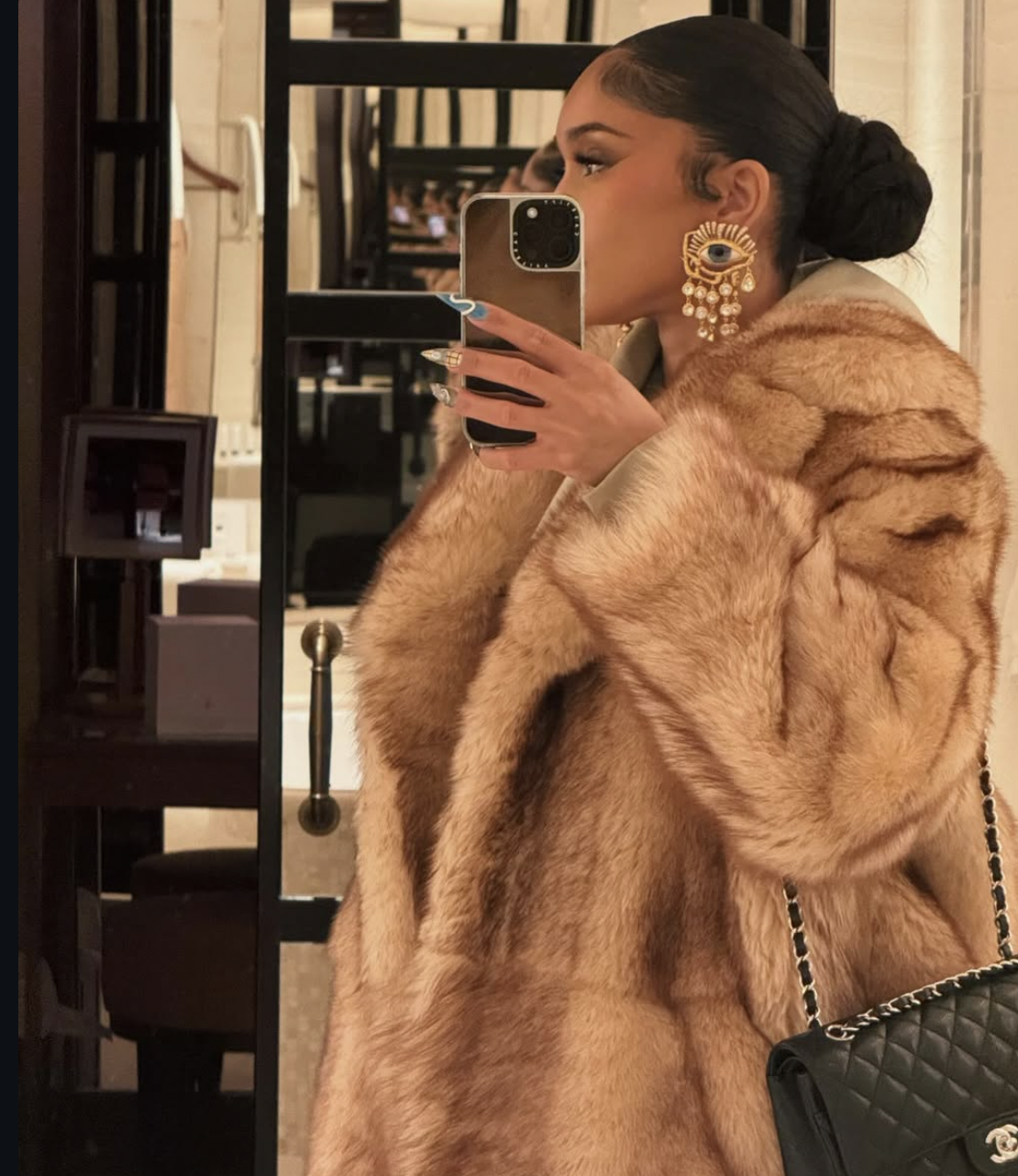 Saweetie taking a mirror selfie while wearing a large tan faux fur coat, showcasing a sleek, high polished bun that adds vertical height to elongate and define a round face shape.