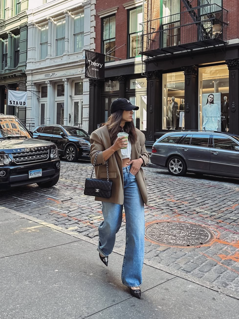 A woman walking down a cobblestone street wearing wide-leg blue jeans, black pointed-toe heels, a tan blazer, and carrying a black quilted Chanel shoulder bag.