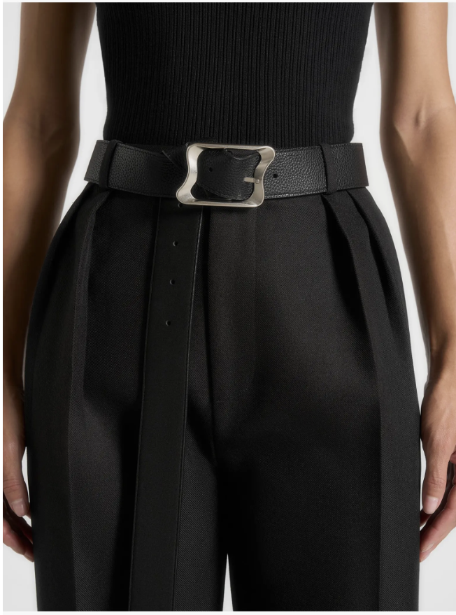 A close-up of a wide black textured leather belt featuring a large, asymmetrical silver statement buckle, styled over black trousers.