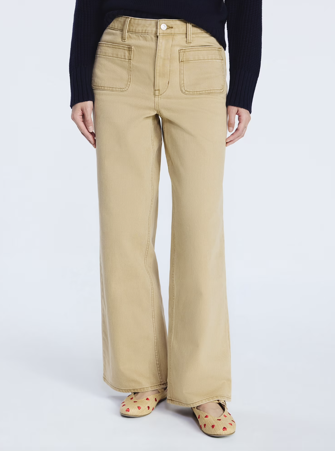 A close-up of a person wearing Free Assembly high-waisted wide-leg jeans in a khaki brown tint, featuring large symmetrical front patch pockets and a gold-tone button closure.