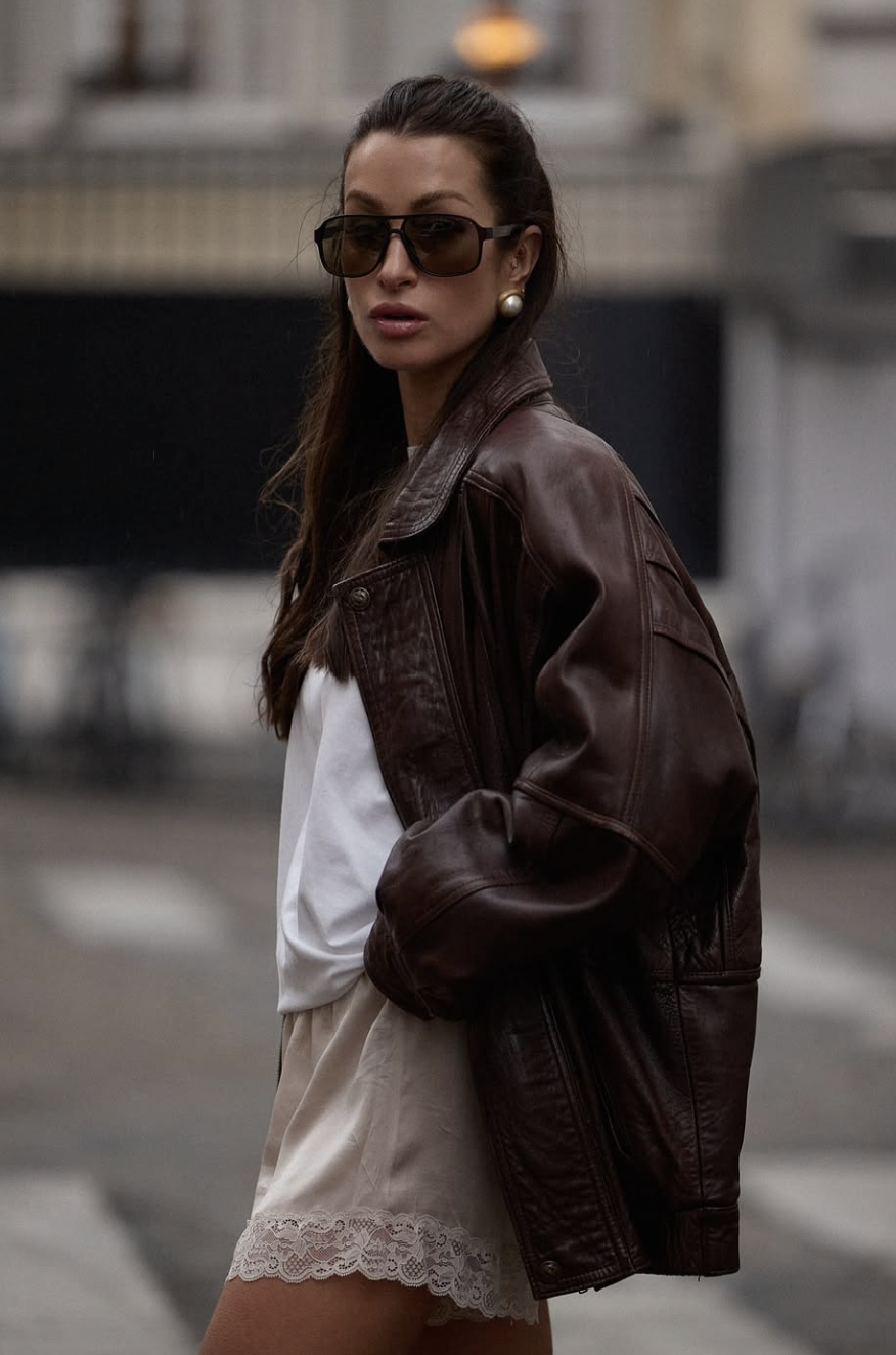 A close-up of a woman wearing cream-colored satin silk shorts with a wide white lace hem, paired with an oversized dark brown leather jacket.