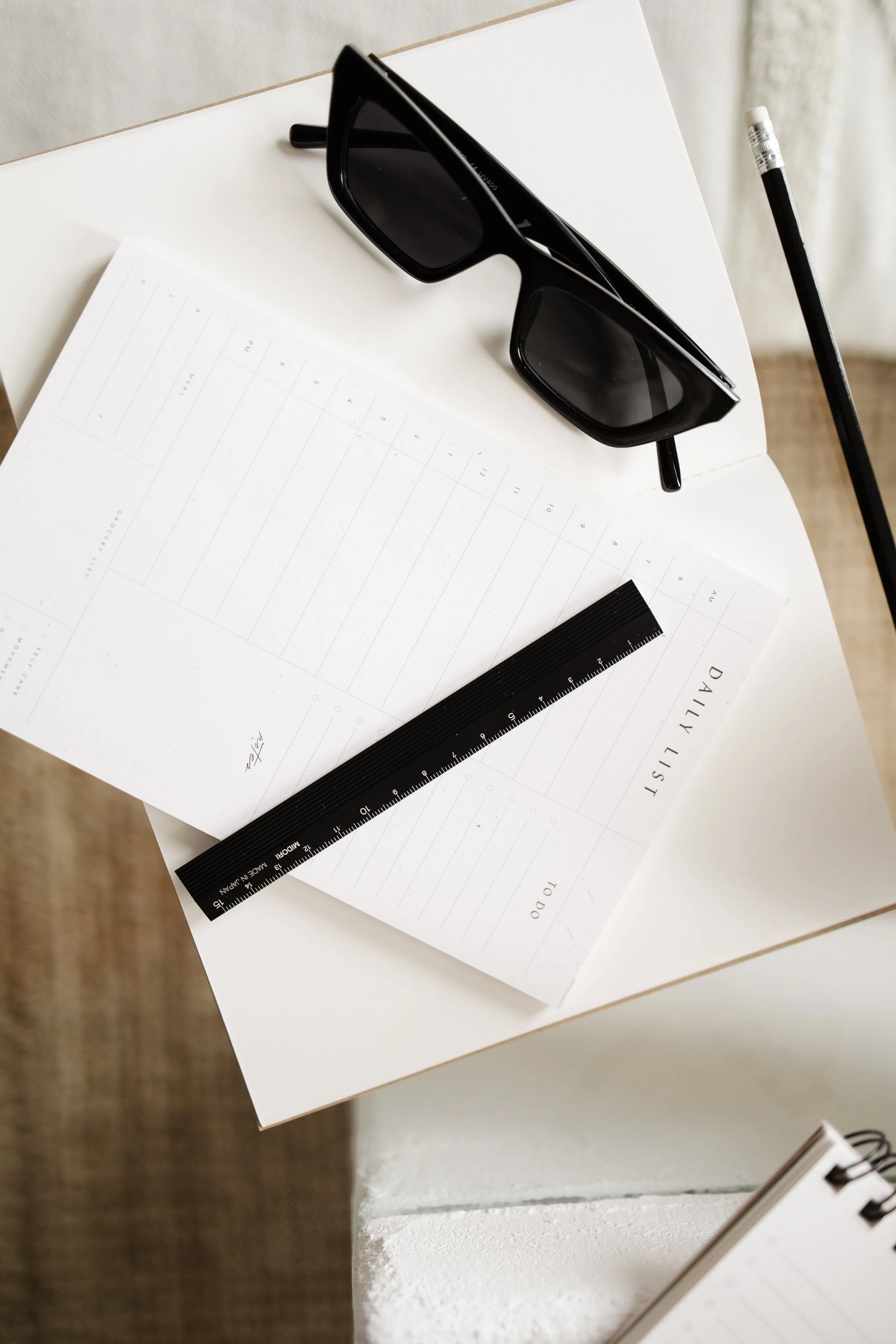 Black sunglasses, a black pencil, a black ruler, and a white daily planner with blank pages on a white surface.