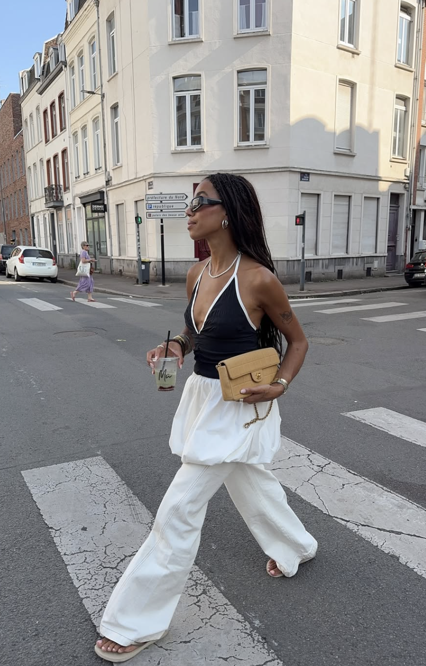 A woman walking across a city street wearing an elevated summer outfit featuring a black halter top layered over a white strapless bubble hem top, paired with relaxed white wide-leg trousers and a tan quilted leather shoulder bag.